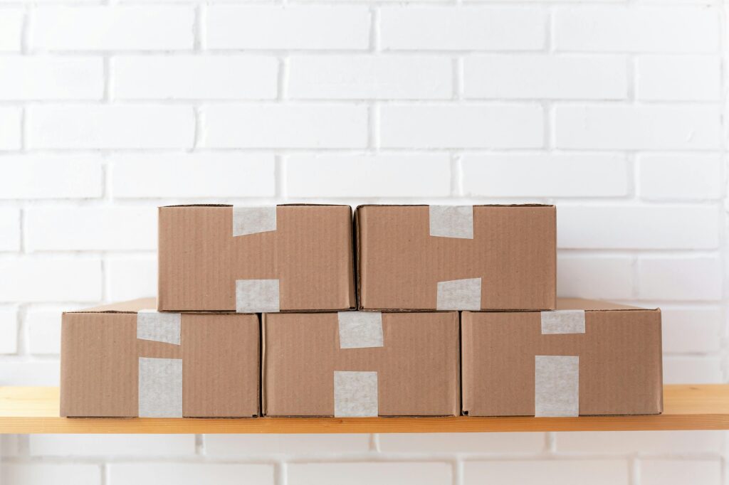 A neat stack of cardboard boxes taped and placed on a wooden shelf against a white brick wall.